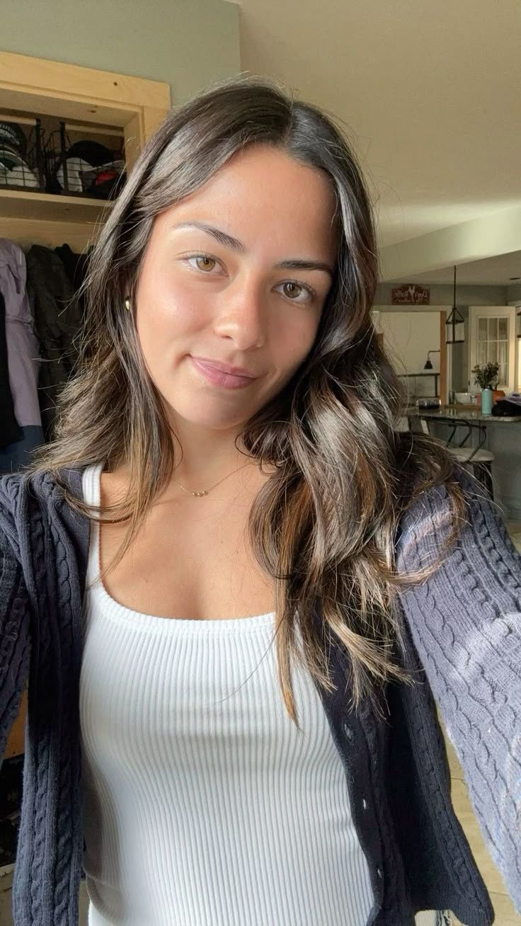Woman with long, wavy hair wearing a white top and dark cardigan indoors.